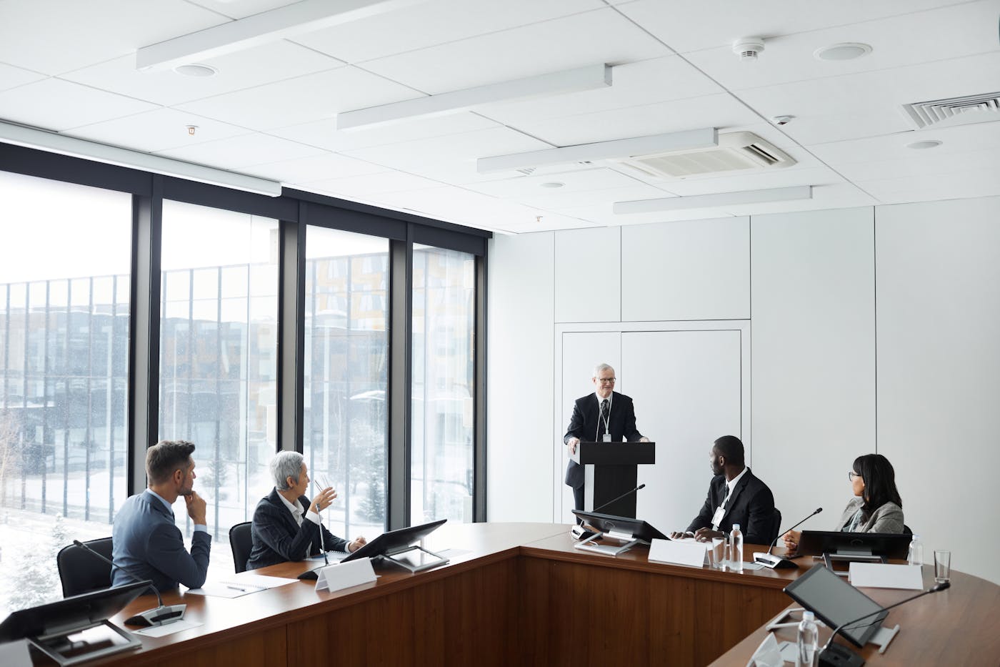 Formal boardroom briefing with a speaker addressing seated executives at a wood conference table.