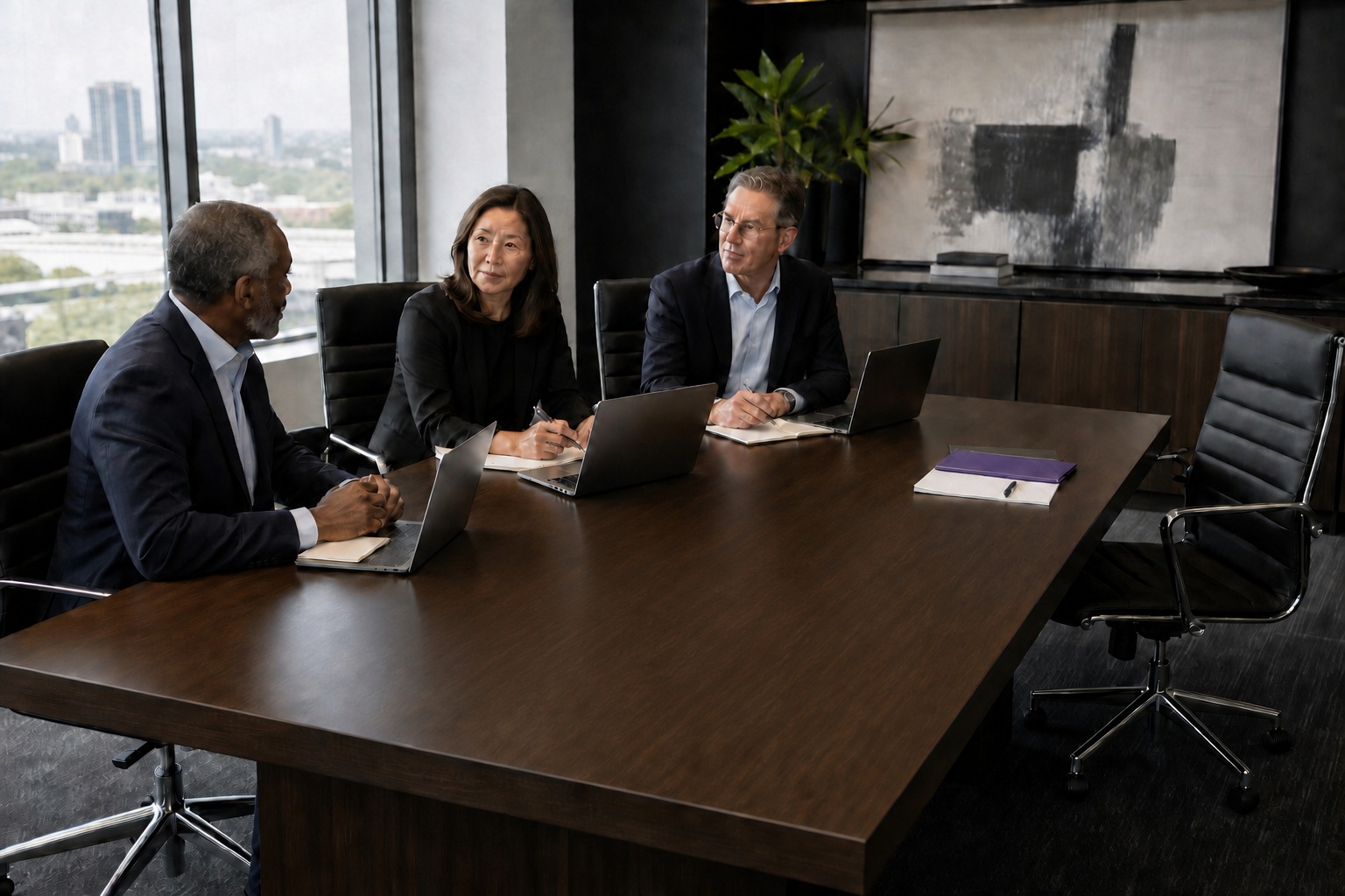 Three executives discuss business strategy around a rectangular boardroom table with laptops and one open seat.